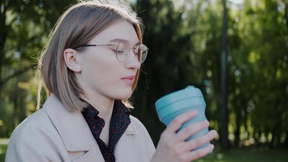 Girl Sits on a Park Bench in Autumn Drinking From an Eco Cup and Looking Around alt