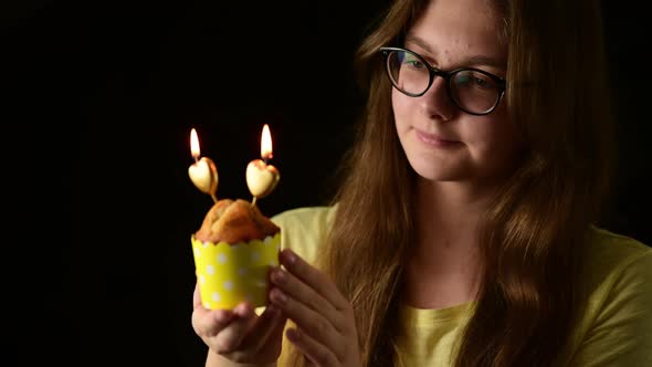 Happy attractive young woman holding one muffin with two heart shape candle, blows out the candles alt