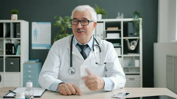 Portrait of Happy Doctor in White Gown Talking and Waving Hand Looking at Camera in Office alt