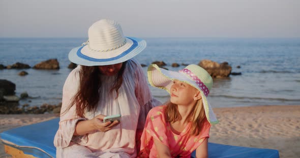 Mom and Daughter on the Beach are Looking in Smartphones Smiling and Talking