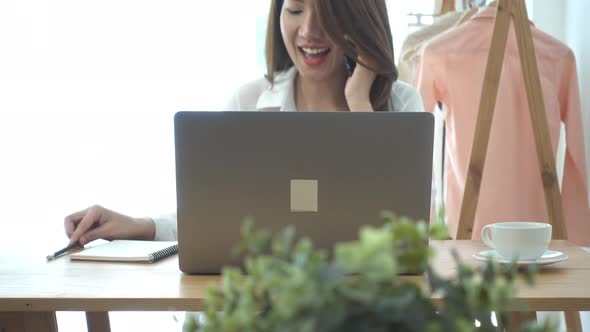 young smiling asian woman working on laptop while sitting in a living room at home using phone. alt