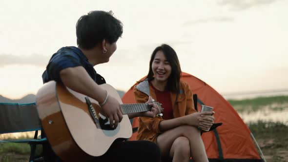 A happy young Asian couple backpacker playing the guitar and singing a song together beside camping.