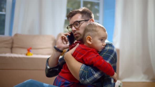 Caucasian Man Holding Toddler Son While Talking on Phone Sitting on Floor at Home alt