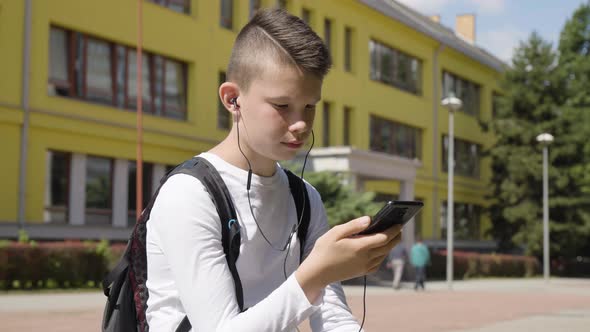 A Caucasian Teenage Boy Listens to Music with Earphones on a Smartphone  a School in the Background