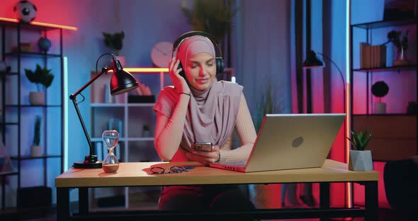 Muslim Woman in Headscarf Enjoying Favourite Music in Headphones from Phone Playlist at home alt