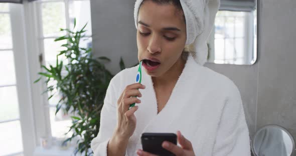 Mixed race woman brushing her teeth and using her smartphone in bathroom alt
