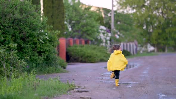 Girl in Yellow Raincoat and Boots has fun and Runs through the Puddles on the Road.
