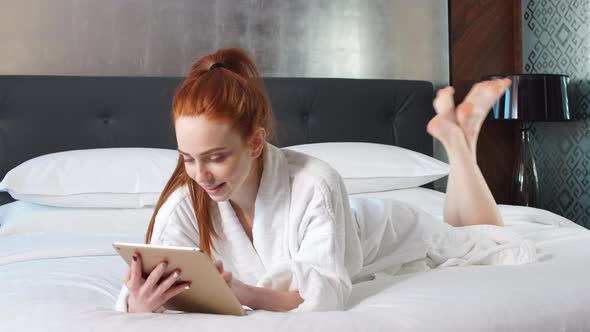 Young Redhead Woman Using Tablet Computer Lying on Bed in Hotel Room