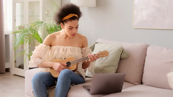 Young african american woman taking online ukulele music guitar lessons tutorial on laptop alt
