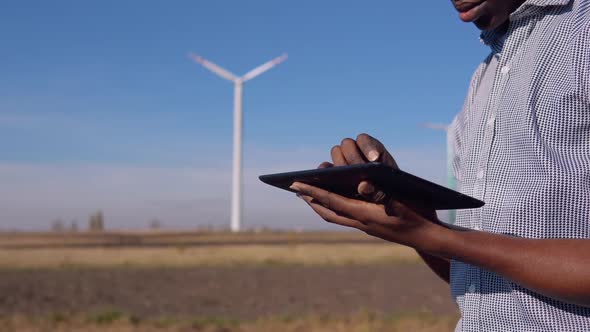 An AfricanAmerican Electrician in a Helmet Stands Against the Backdrop of a Windmill at an Air Power alt