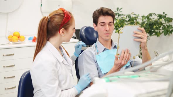 Female Dentist Holding Mirror and Showing Teeth Problems to Male Patient in Dental Office alt