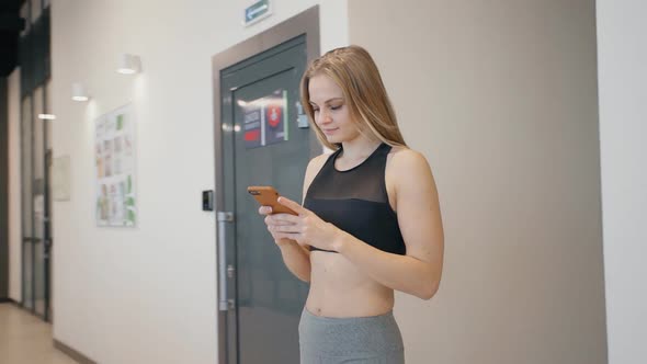 Hipster Young Woman Using Smart Phone To Surf Internet in Gym After Workout. Smiling Girl Chats.