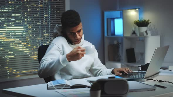 Mixed-race Man Working on Computer, Speaking By Smartphone and Drinking a Cup of Tea alt