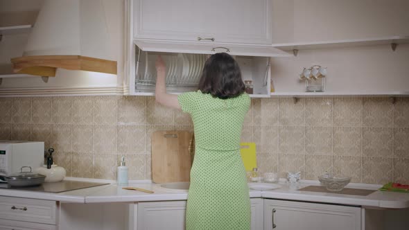 Brunette Girl in a Green Dress Puts Things in Order in the Kitchen Cabinet Arranges the Dishes alt