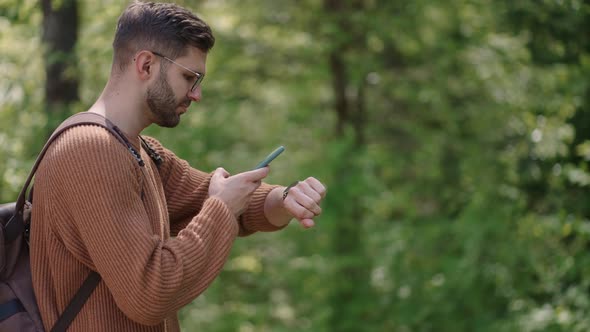 A Young Bearded Man with Glasses with a Backpack Adjusts a Smart Watch Using His Phone to alt