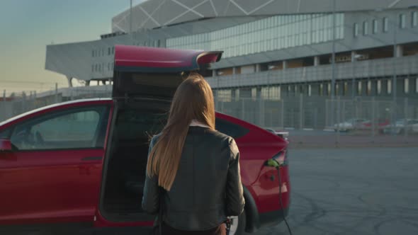 Close Up of a Girl Approaches an Electric Car Which is Charging at the Station alt