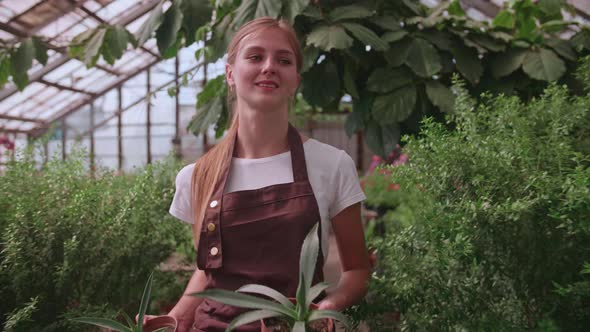 Young Blonde Girl Walks Through a Greenhouse with Pots of Flowers
