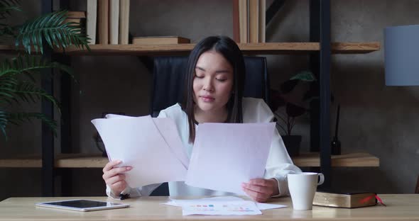 Asian Woman Indoor Sitting in the Workplace While Looking Through Paper Documents in Loft Apartment alt