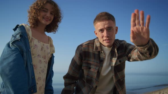 Happy Couple Waving Hands Saying Hello on Camera Standing Sandy Beach alt