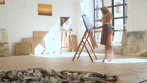 Woman Artist Paints on Canvas on Easel and Holds Palette in Art Workshop alt