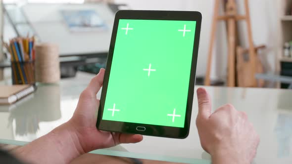 Man Holds a Black Tablet with Green Screen on in Both Hands and Keeps Watching alt
