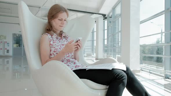 Attractive Businesswoman Sitting on a White Chair in a Glass Office and Checks Documents