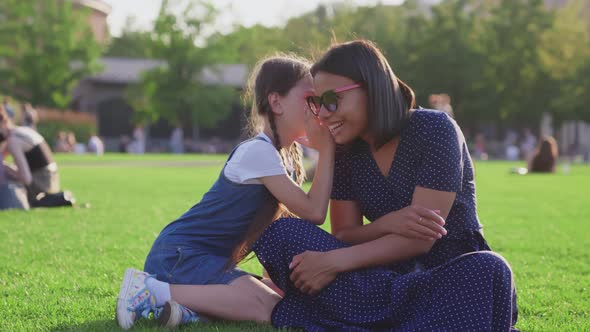 Portrait of Caucasian Girl Telling African Mother Secret Sitting Together on Lawn in Park alt