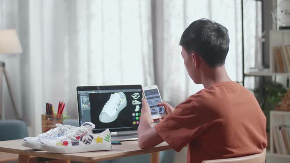 Asian Boy Footwear Designer Looking At Photos On Smartphone While Designing Shoe On A Laptop alt