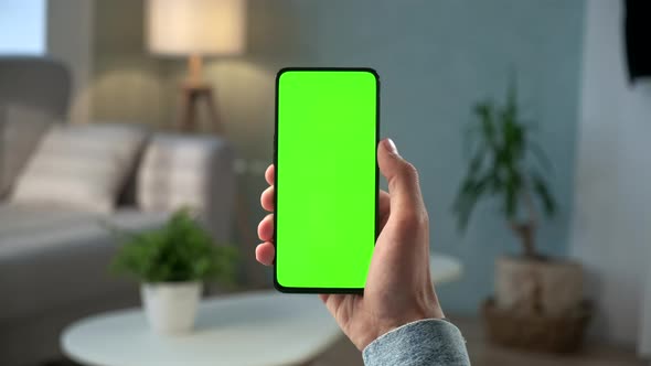 Close-up Shot of Young Man at Home Sitting on a Chair Using With Green Mock-up Screen Smartphone alt