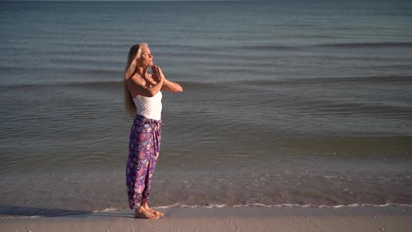 Slow motion of mature woman in sarong in a prayer pose on a beach at sunrise or sunset. alt