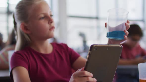 Caucasian schoolgirl using tablet looking at blue liquid in jar during chemistry class alt