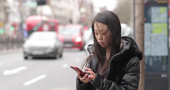 Chinese woman waiting at bus stop, using smartphone alt