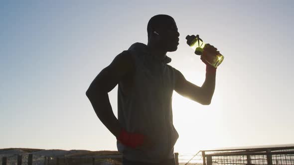 African american man drinking water, taking break in exercise outdoors by the sea alt