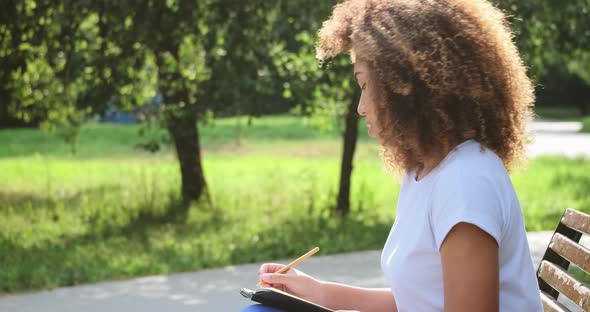 Pensive Mixed Race Female Handwriting Paper Notes in Notepad Page Use Pencil at Park alt