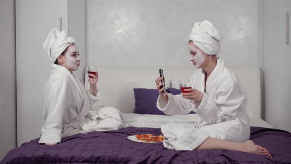 Young Girls in Robes with Towels on Their Heads Sit on the Bed Drink Wine