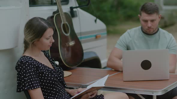 Traveler Man working at laptop and woman reading the book in summer camping. alt