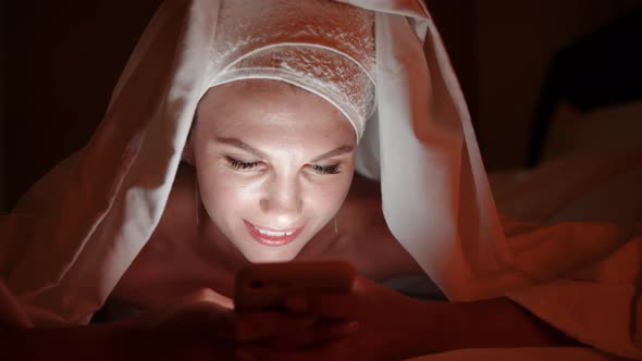 Beautiful Young Woman Using Smart Phone Under Blanket in Bedroom on Bedtime alt