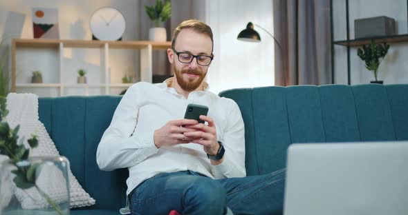 Man in Glasses Sitting in Relaxed Pose on Soft Couch at Home and Browsing Phone Apps alt