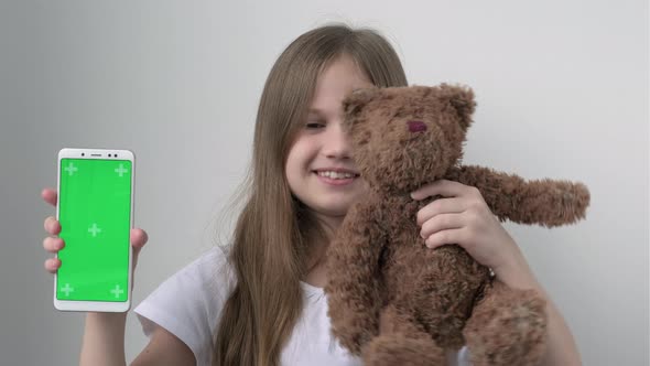 Cute Child Girl Shows Phone with Green Screen Mock Up and Hugs Teddy Bear alt