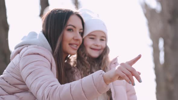 Young Pretty Mother Is Pointing at Something Hugging Her Daughter in a Park alt