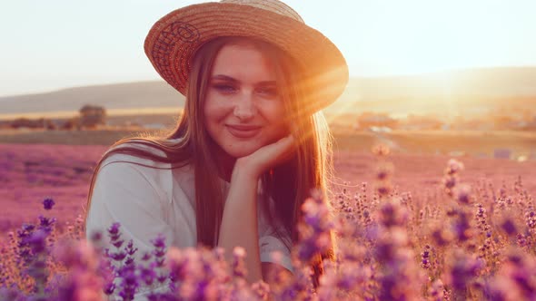 Beautiful Young Girl in a Straw Hat and White Dress Sitting in a Lavender Field on Sunset