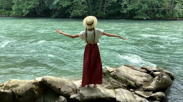Young beautiful vintage woman in long skirt and straw hat dancing on stones near mountain river