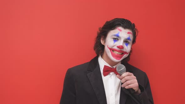 A Clown with Bright Makeup on His Face in a Suit Stands with a Microphone in His Hands on an