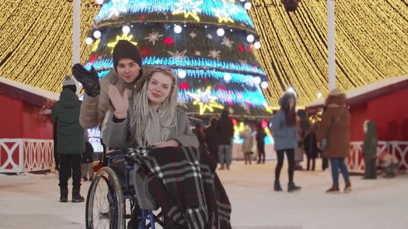 Smiling Young Woman in a Wheelchair and Her Boyfriend at Christmas Party on the Streets Waving Hands alt