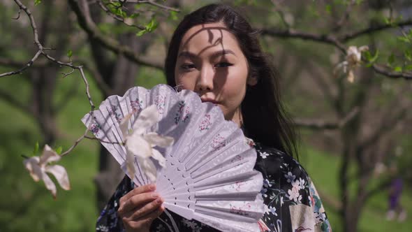 Live Camera Approaches to Confident Asian Beautiful Woman Holding Fan Standing in Blooming Spring alt