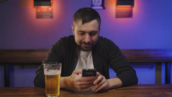 Portrait of Man with Glas of Beer Looking at Smart Phone in Pub 