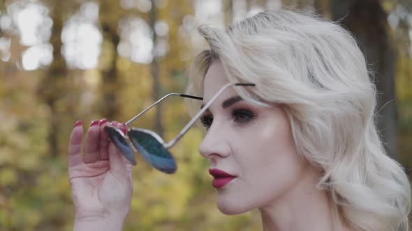 Seductive Blonde with Red Lips Puts on Sunglasses and Looks Straight in Park alt