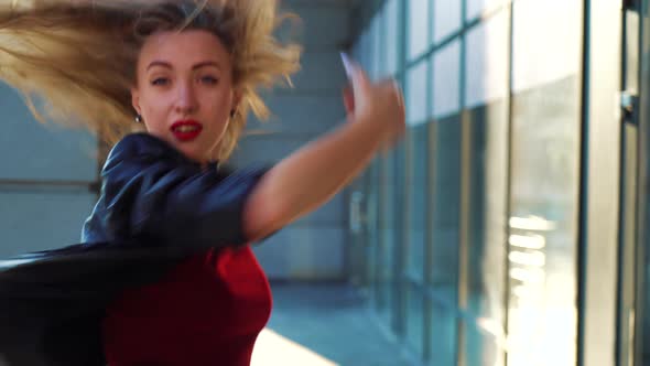 Young Blonde Woman Looks in the Camera and Dances in Rays of Sunset on Street alt