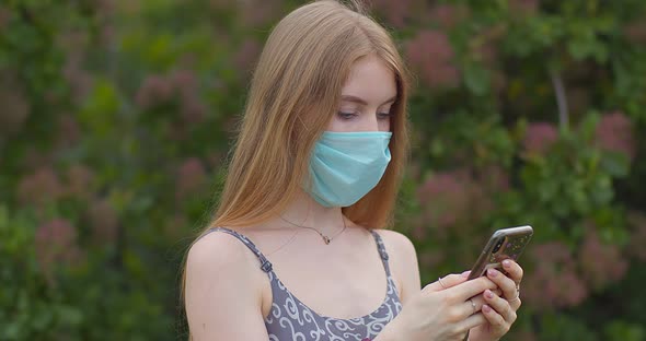 A young girl in a protective medical mask looks at the phone and sends messages