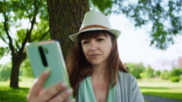 Brunette Woman in a Hat Sits in the Park Under a Tree and Uses the Phone